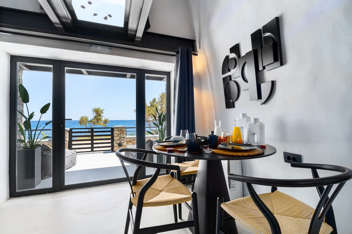 A dining area features a round table set for breakfast, complemented by woven chairs. Large glass doors open to a private balcony with a sea view, bringing natural light into the space. Coastal decor elements enhance the modern design.