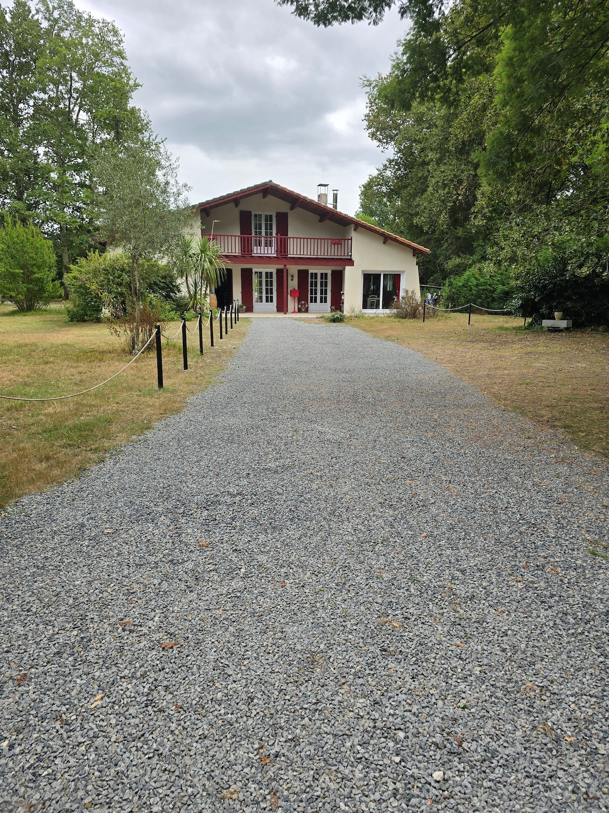 A gravel driveway leads to a two-story house surrounded by lush green landscaping. Large windows and a red front door are prominent features. The area shows ample outdoor space, with trees and shrubs providing a natural setting.
