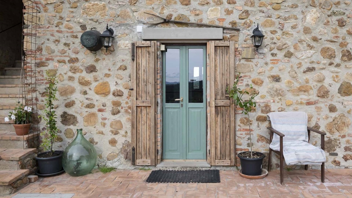 An inviting entrance features a green double door, framed by wooden shutters. A stone wall adds character, while a rustic chair sits nearby. Potted plants and a decorative glass object enhance the outdoor space, creating a welcoming atmosphere for guests.