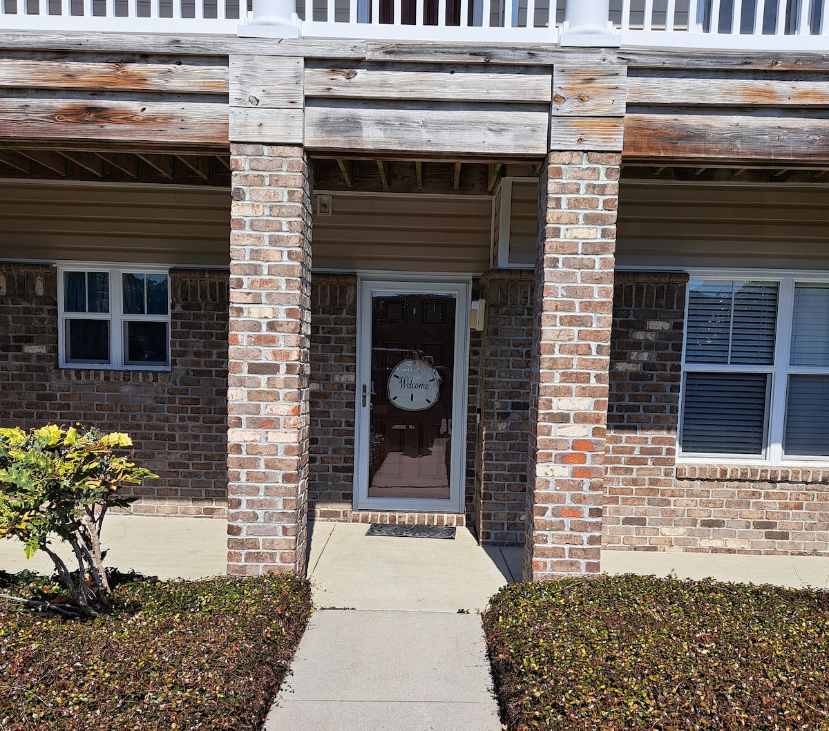 The entrance to the unit is framed by brick pillars and features a welcoming door with a decorative sign. Lush green shrubs line the pathway, and the concrete walk is well-defined, leading up to the door. Two windows provide natural light to the area.