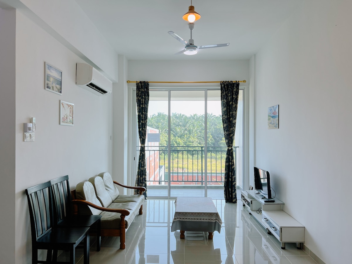 A bright and airy living room features a comfortable sofa set and a small coffee table. A flat-screen TV is positioned on a sleek white media unit. Large glass doors allow ample natural light and provide a view of the lush greenery outside.