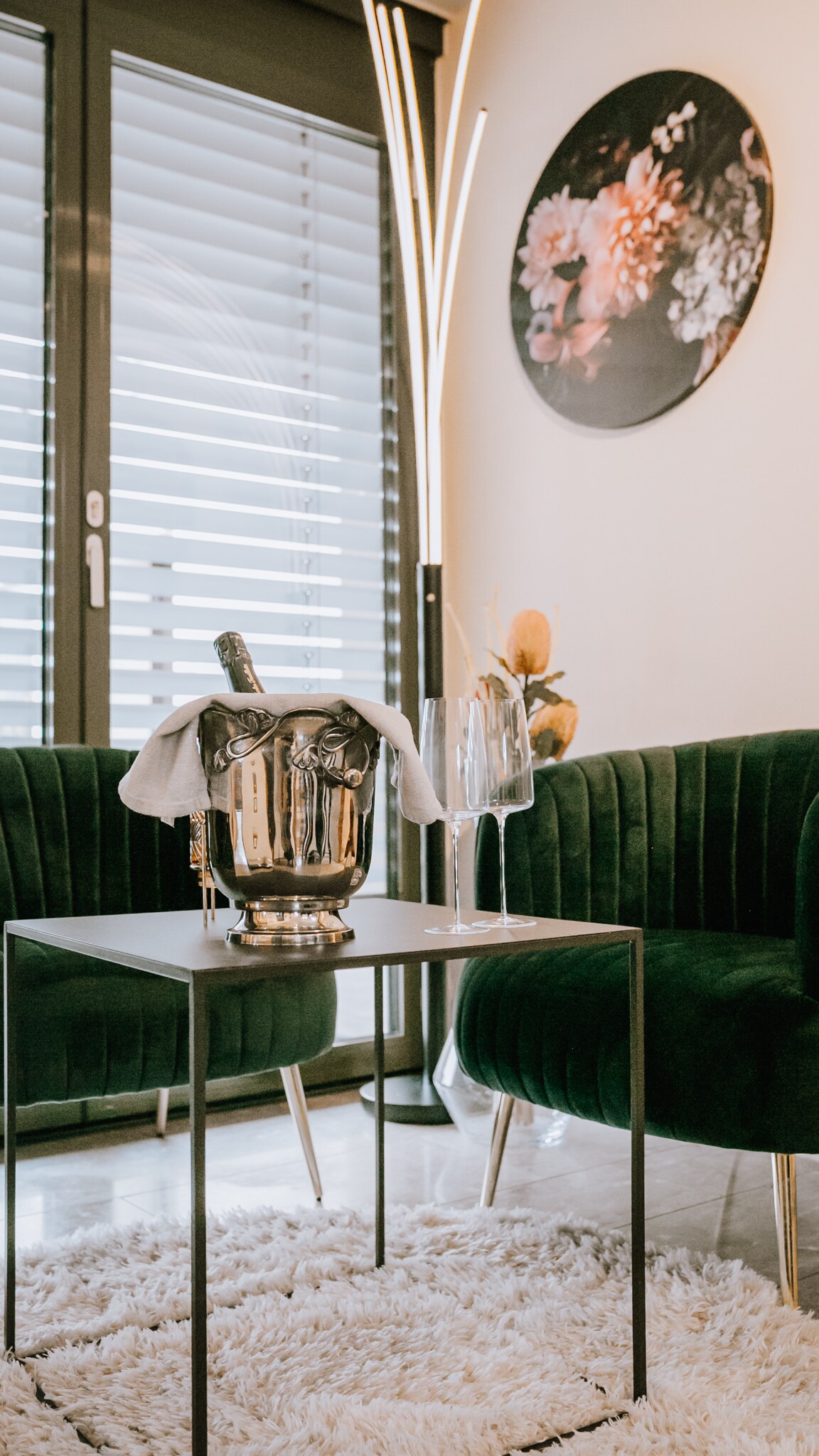 A modern sitting area is showcased with two green velvet armchairs positioned around a sleek black table. An ice bucket sits on the table, covered with a towel, alongside two crystal glasses. Natural light enters through the large glass doors, enhancing the warm ambiance.