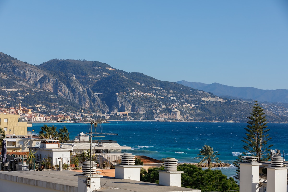 A view of the coastline showcases rolling hills and the blue sea extending to the horizon. The scene is complemented by green palm trees and terraced rooftops in the foreground, while a clear sky adds to the serene atmosphere.
