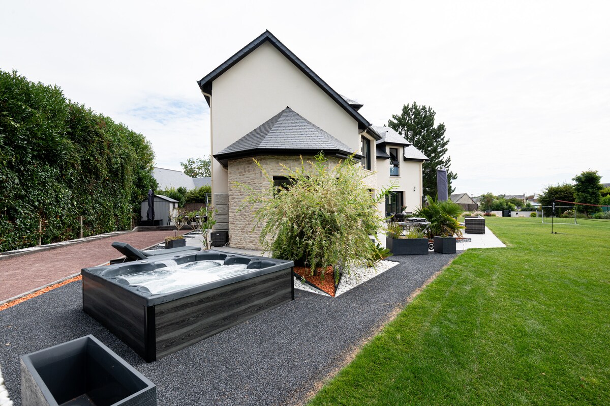 A sleek and modern spa is positioned on a gravel path surrounded by landscaped greenery. The exterior of the house features light-colored walls and a multi-pitched roof, with large windows framing the inviting backyard, which is set against an expansive lawn.