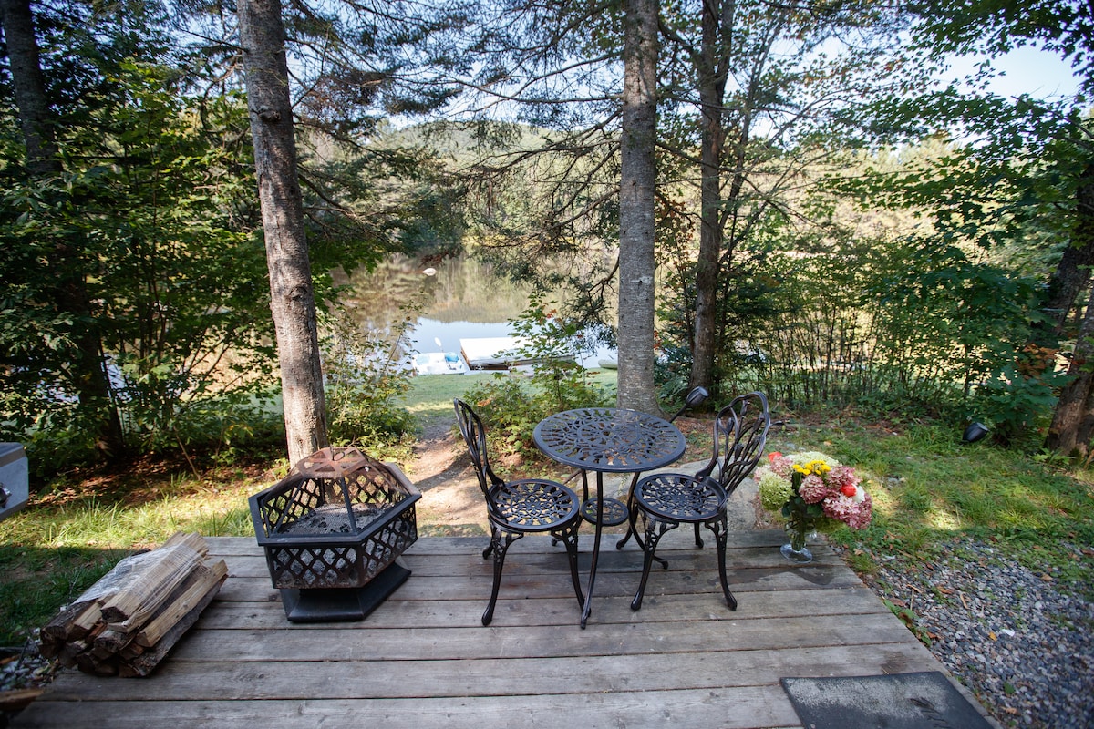 A private deck offers a round table with three elegant chairs, positioned nearby a fire pit. A bouquet of flowers adds a touch of color. Through the trees, a serene pond is visible, providing a peaceful backdrop to this outdoor space.