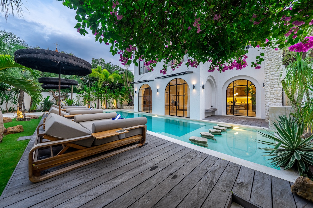A serene outdoor space is centered around a tranquil swimming pool, surrounded by lush greenery. Sun loungers with shaded umbrellas are positioned beside the water, while stepping stones lead across the pool. The villa’s elegant architecture features arched windows and a textured stone wall.