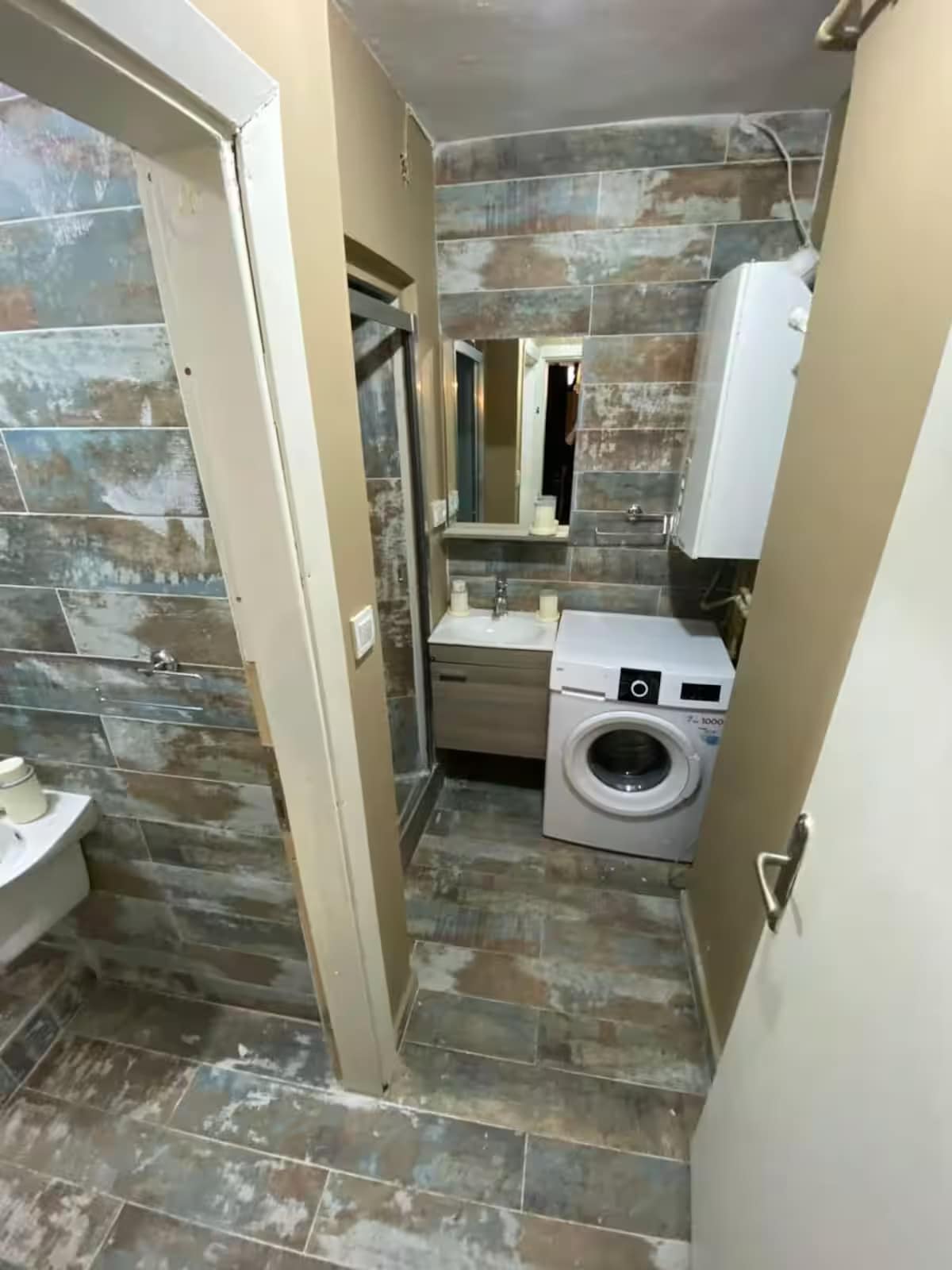 A shared bathroom is shown featuring a narrow design with earthy-toned walls and stone tiles. A washing machine sits next to a compact sink and a mirror. A shower area is visible, connected by an open space leading to the entrance.