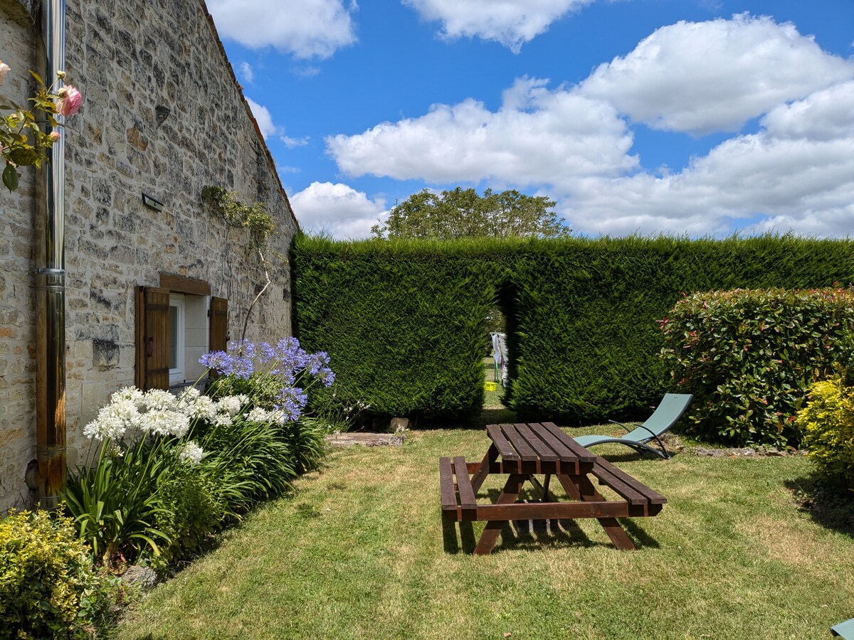 A garden area features a grassy lawn surrounded by vibrant flowering plants. A picnic table and a reclining lounge chair are positioned on the lawn. A tall hedge provides privacy, with a clear blue sky and fluffy clouds visible above.