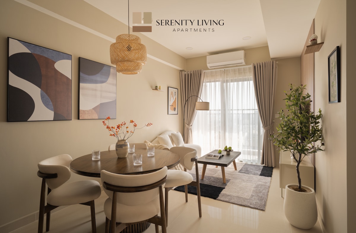 A modern dining area is presented with a round wooden table surrounded by four upholstered chairs. Two abstract wall art pieces and a floor plant add a touch of style. Natural light filters through the large window, while soft curtains create a serene ambiance.