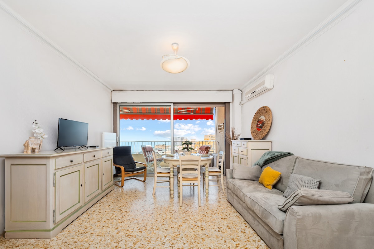 A bright living room is displayed with a cozy sofa and a TV set on a light-colored cabinet. Natural light floods the space through the large balcony doors, which lead to a terrace adorned with outdoor seating. A dining table is positioned nearby, accommodating several guests.