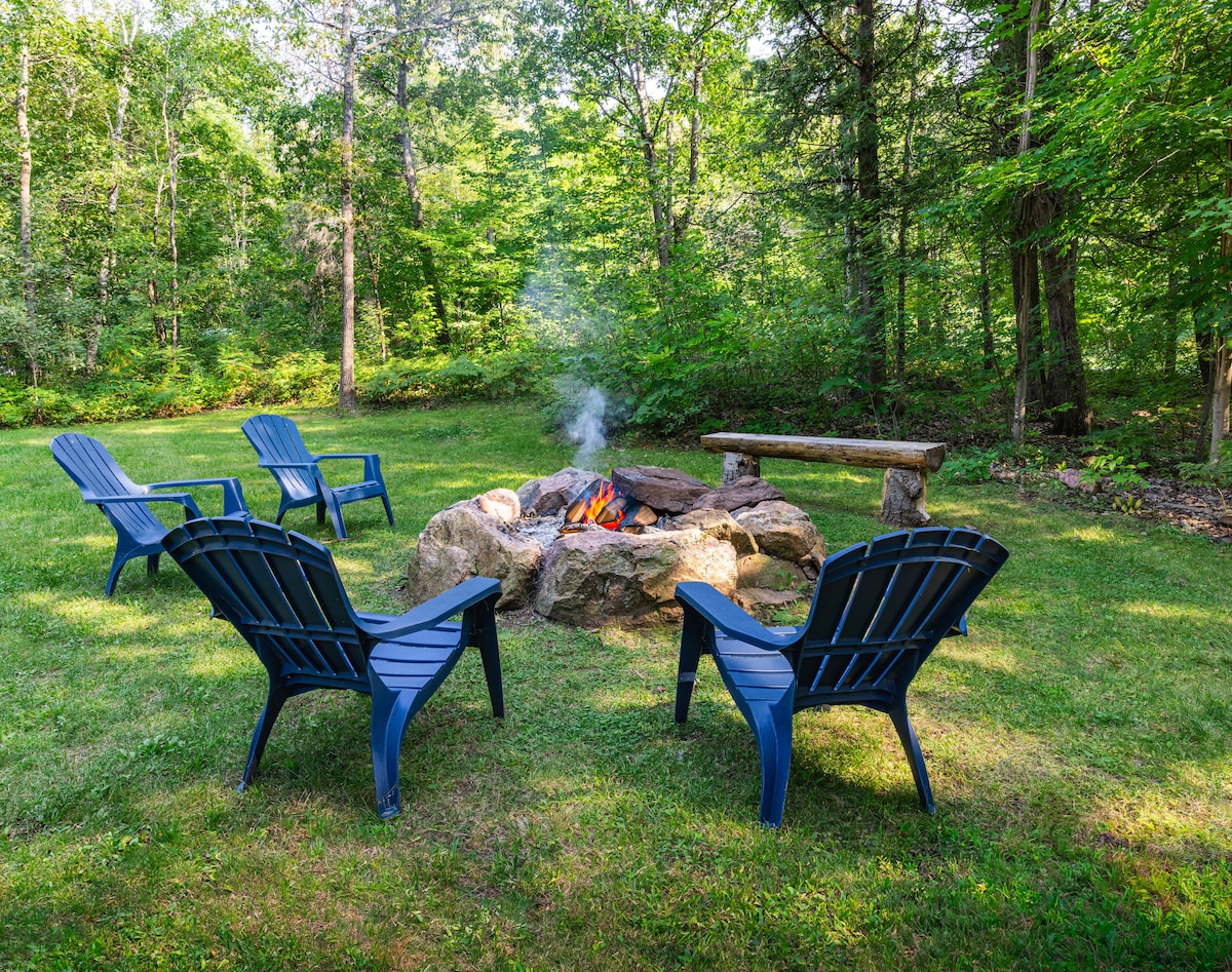 A tranquil outdoor space is surrounded by trees, featuring a fire pit encircled by several blue Adirondack chairs. A rustic wooden bench is positioned nearby, providing a perfect spot to relax. Wisps of smoke rise from the glowing fire, inviting gatherings and warm moments.