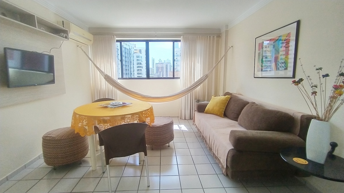 A bright living area features a comfortable sofa and a round table set with a decorative cloth. A hammock is suspended across the room, while large windows allow for natural light and views of the surrounding buildings. A minimalist TV unit is mounted on the wall.