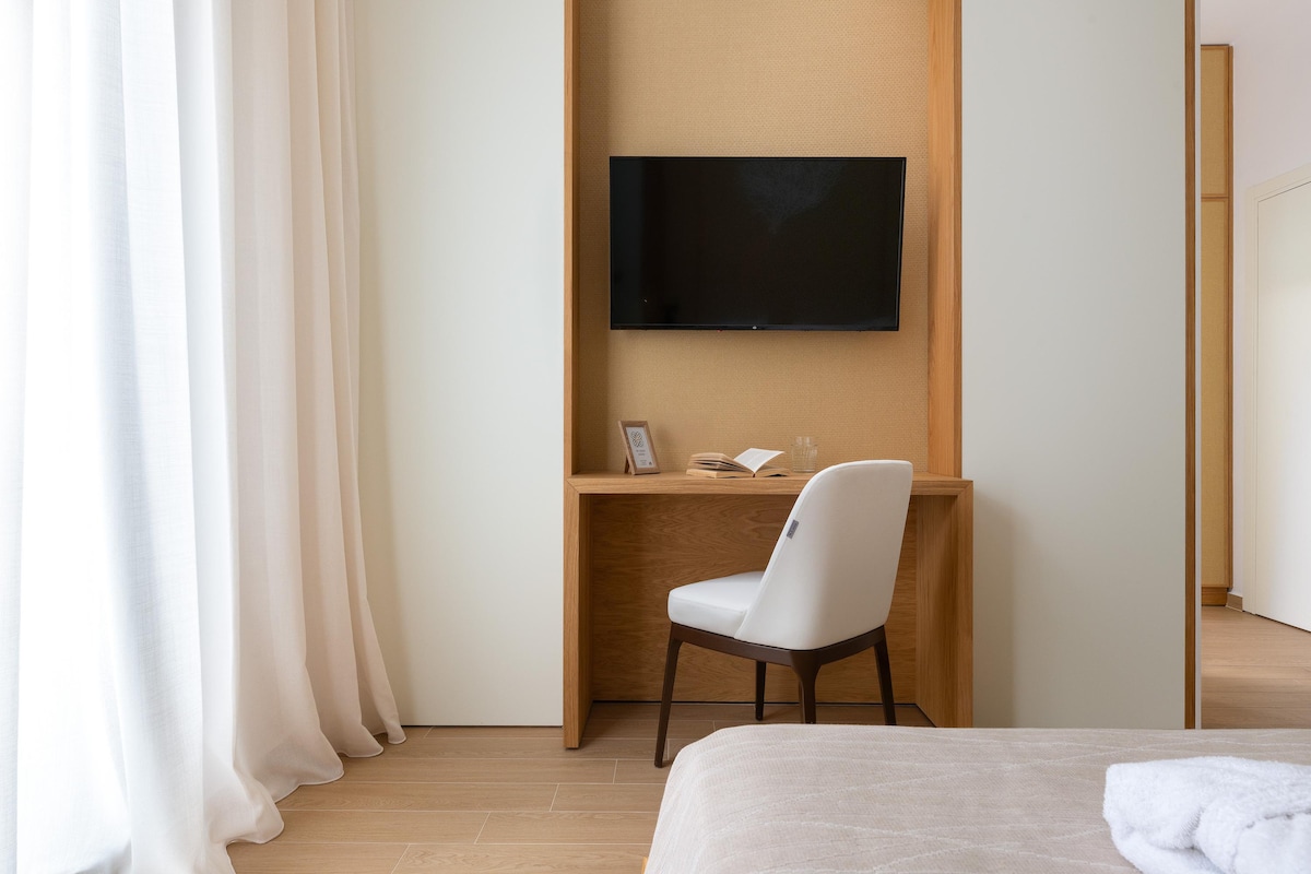 A modern room features a wooden desk and a white chair positioned against a beige wall. A flat-screen TV is mounted above the desk, while softly draped curtains provide natural light, enhancing the spacious atmosphere of the area.