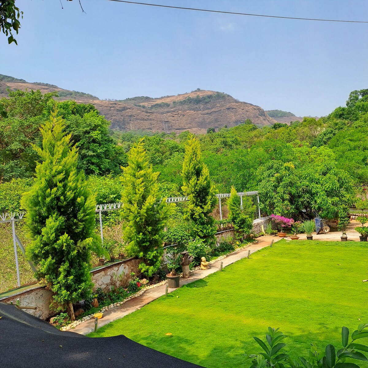 A lush, green garden is presented, featuring neatly trimmed lawns and an array of colorful plants. A backdrop of mountains can be seen in the distance, while neatly lined trees create a serene atmosphere. The sky remains clear, enhancing the natural beauty of the scene.
