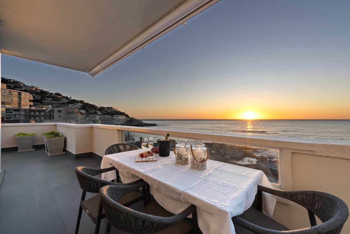 An outdoor dining table is set with a white tablecloth and glassware, facing a vibrant sunset over the Atlantic Ocean. The balcony features comfortable seating and overlooks coastal views, with the horizon meeting the sea in warm hues of orange and yellow.