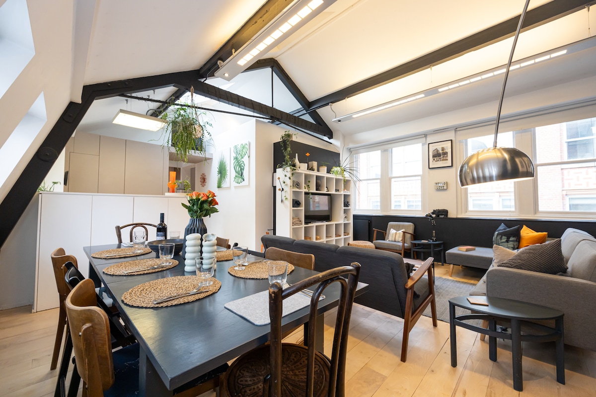 An open-plan living space features a dining area with a large dark table surrounded by mixed seating. A spacious lounge area showcases a cozy sofa and a textured armchair, while natural light floods through large windows and contrasts with the black accent wall.