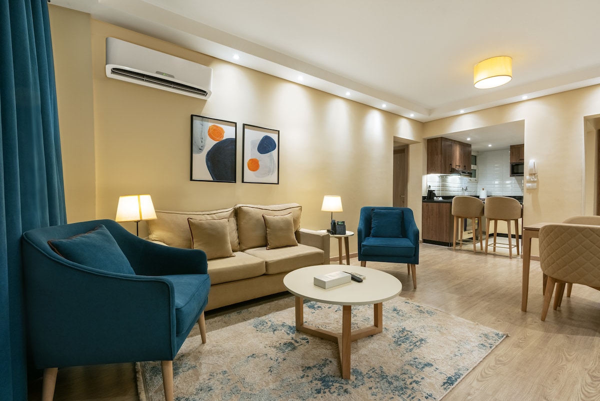 A cozy living area features a comfortable sofa and two accent chairs arranged around a circular coffee table. Soft lighting from two lamps creates a warm ambiance. A rug adds texture to the wooden floor, while modern artwork adorns the wall.