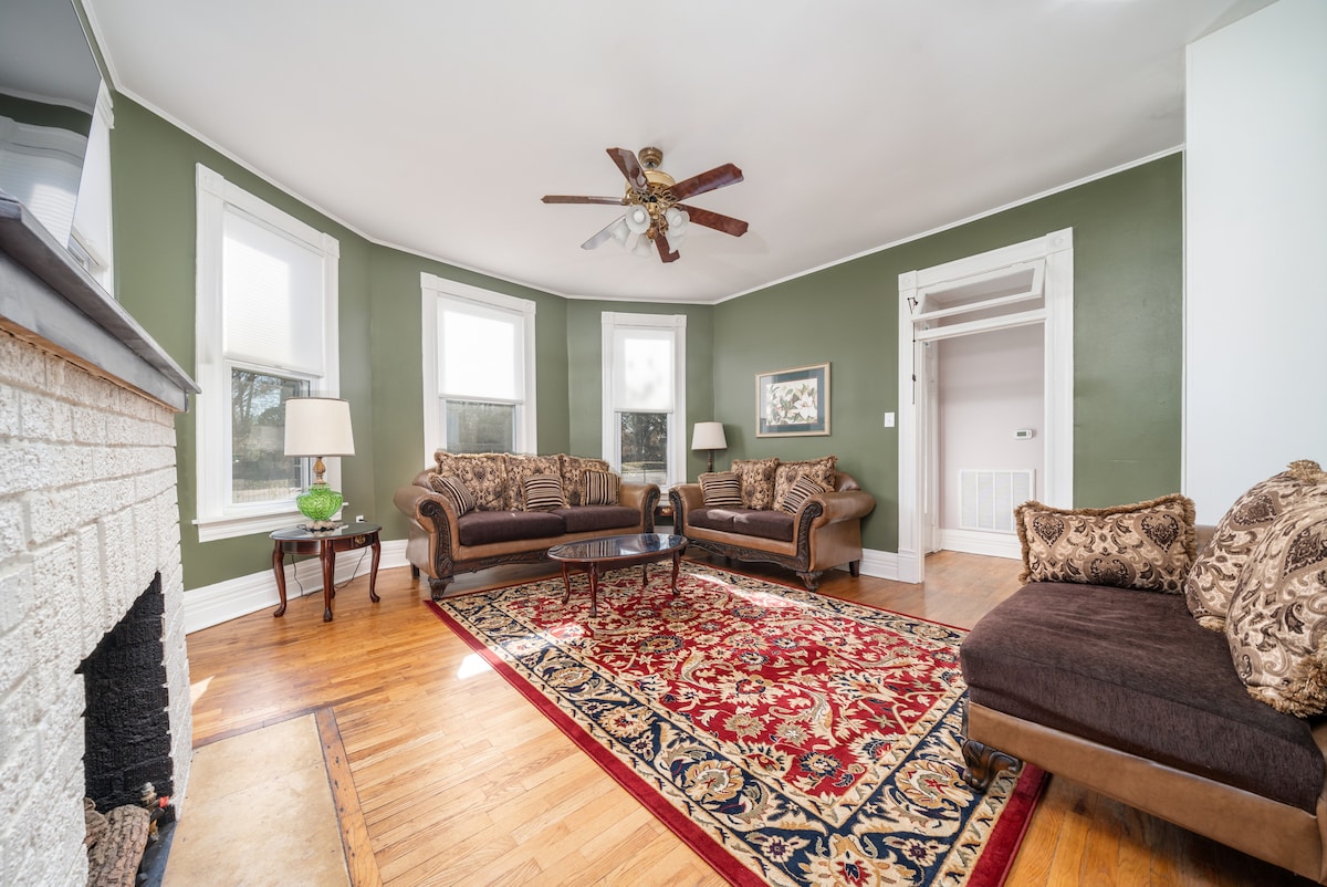 A spacious living area features original hardwood floors and high ceilings. Two comfortable sofas are arranged around a coffee table, with a decorative area rug anchoring the space. Ample natural light fills the room through multiple windows, creating a cozy and inviting setting.