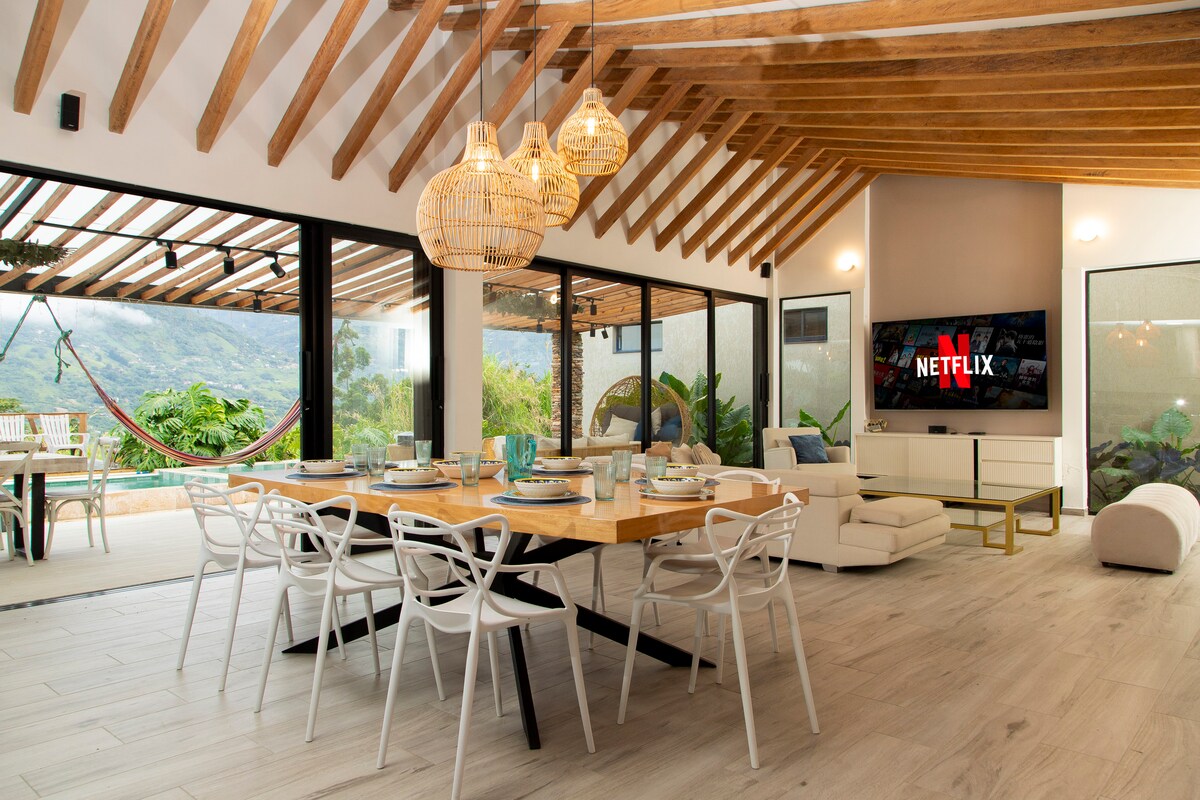 A spacious living and dining area showcases natural wood beams overhead and large glass doors that open to a scenic outdoor space. A modern dining table is set for several guests, while a comfortable seating arrangement includes a stylish sofa. A large television is mounted on the wall.