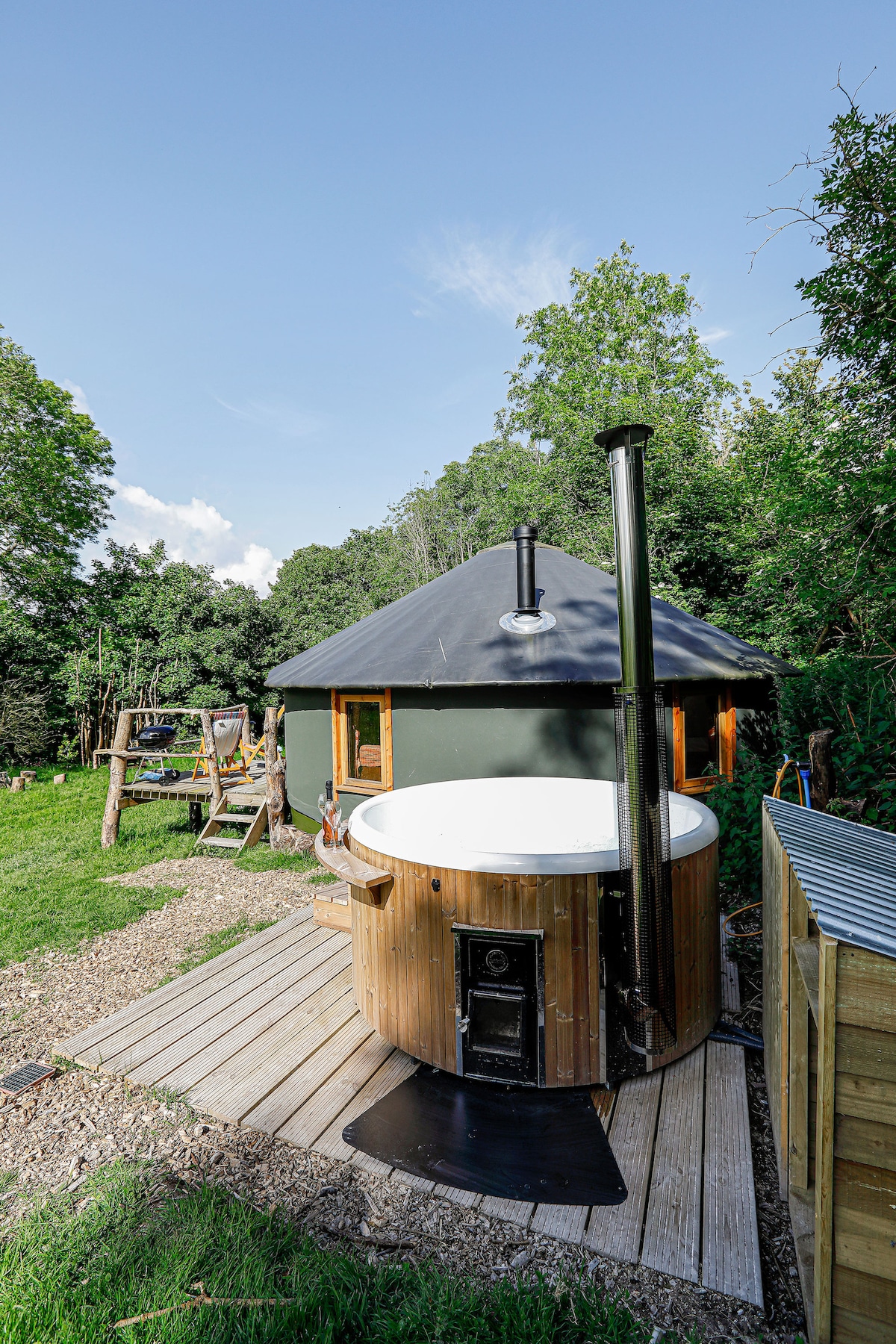 An outdoor view of the yurt is presented alongside a wooden hot tub. The hot tub features a cylindrical chimney and is positioned on a wooden deck, surrounded by greenery. The yurt's gray roof contrasts with the vibrant foliage in the background.
