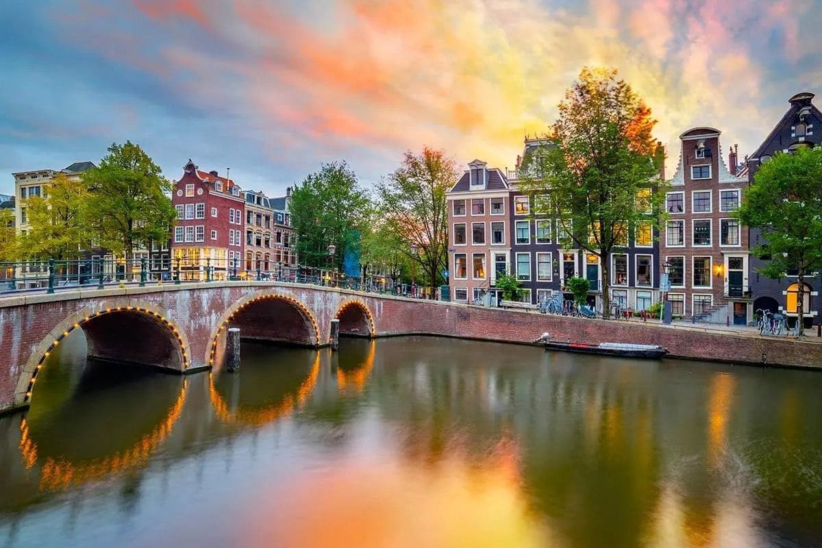 A vibrant sunset is reflected in the calm waters of a canal, framed by classic architecture. Historic buildings with large windows and trees line the bank, while a stone bridge arches gracefully over the water, contributing to the scenic view.