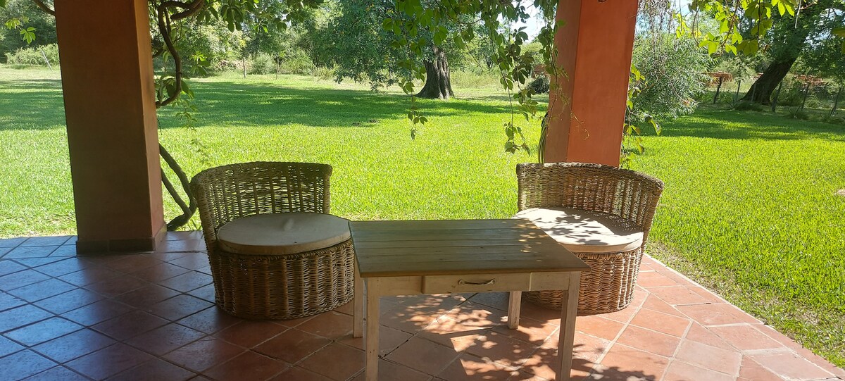 A shaded outdoor seating area features two wicker chairs and a small wooden table. The space is surrounded by lush green grass and trees, creating a tranquil setting for relaxation.