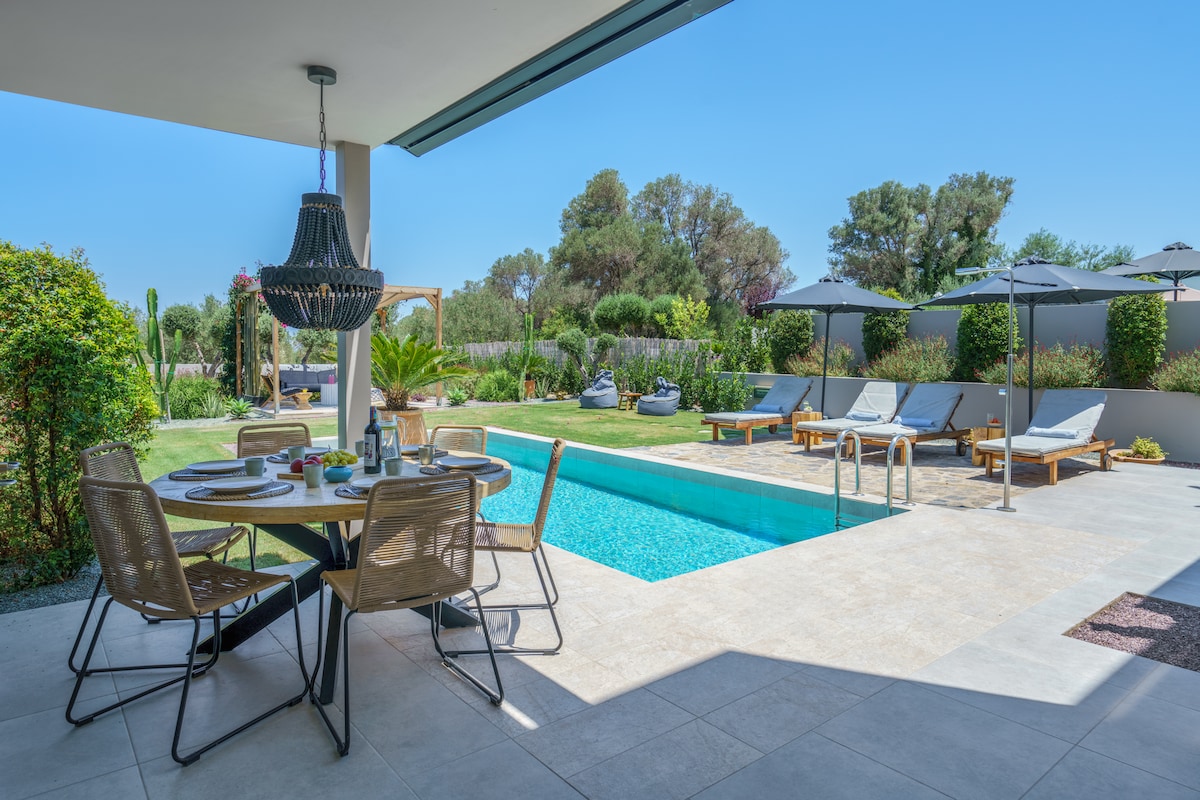 A spacious outdoor dining area is set beside a private swimming pool surrounded by lounge chairs. Lush greenery, including palm and shrubbery, provides privacy. A shaded table with elegant seating accommodates guests, with the inviting blue water of the pool adding a refreshing touch.