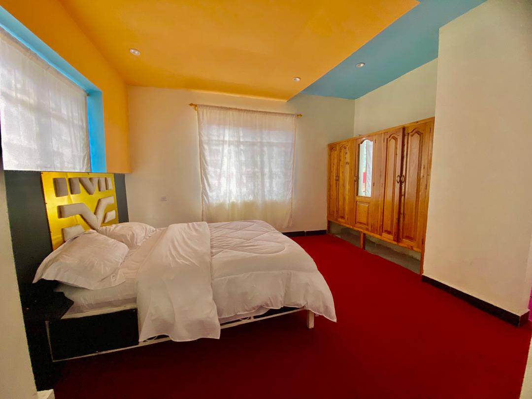 A spacious bedroom features a brightly painted ceiling in light blue, complemented by warm yellow walls. Natural light filters through sheer curtains, illuminating a large bed with white linens. A wooden wardrobe stands against one wall, adding a touch of warmth to the space.