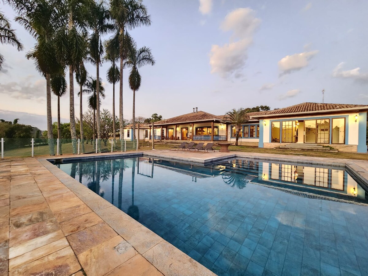 An expansive pool reflects the sky and surrounding palm trees, leading to a spacious outdoor area. The large house, designed with a blend of traditional and modern styles, features multiple windows and seating arrangements for relaxation and gatherings.
