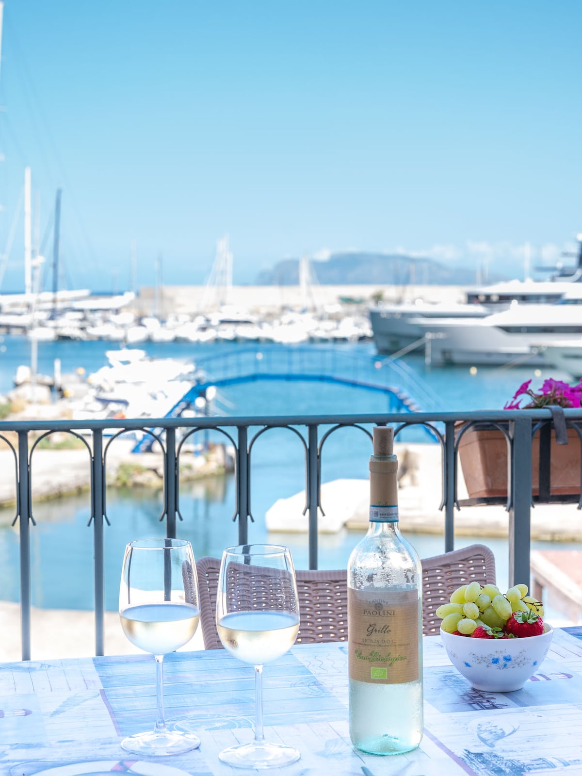 A table set for two is adorned with two glasses of white wine and a bottle, accompanied by a bowl of grapes. The backdrop reveals a scenic view of a marina with multiple boats and a bright blue sky.