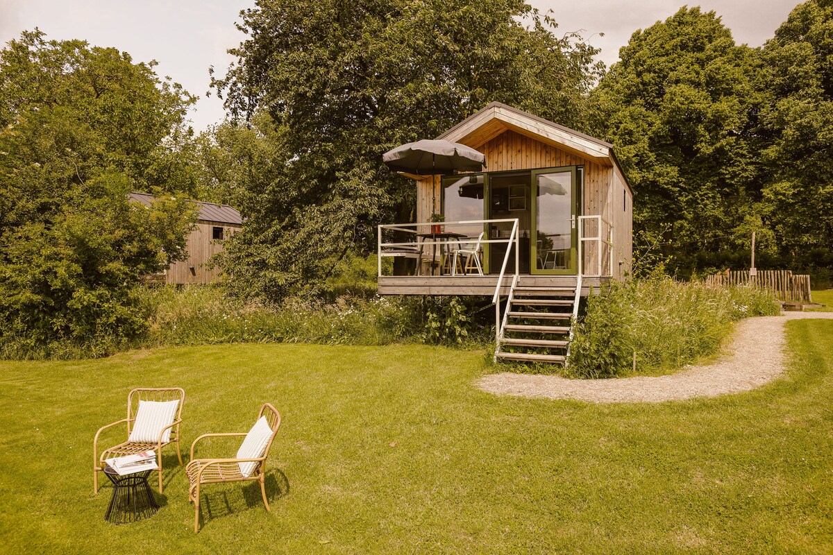 A wooden cabin is framed by lush greenery and set on a grassy area. Two rattan chairs are positioned on the deck, accompanied by a table and umbrella, creating a cozy outdoor seating space. A winding path leads to the cabin, enhancing the natural surroundings.