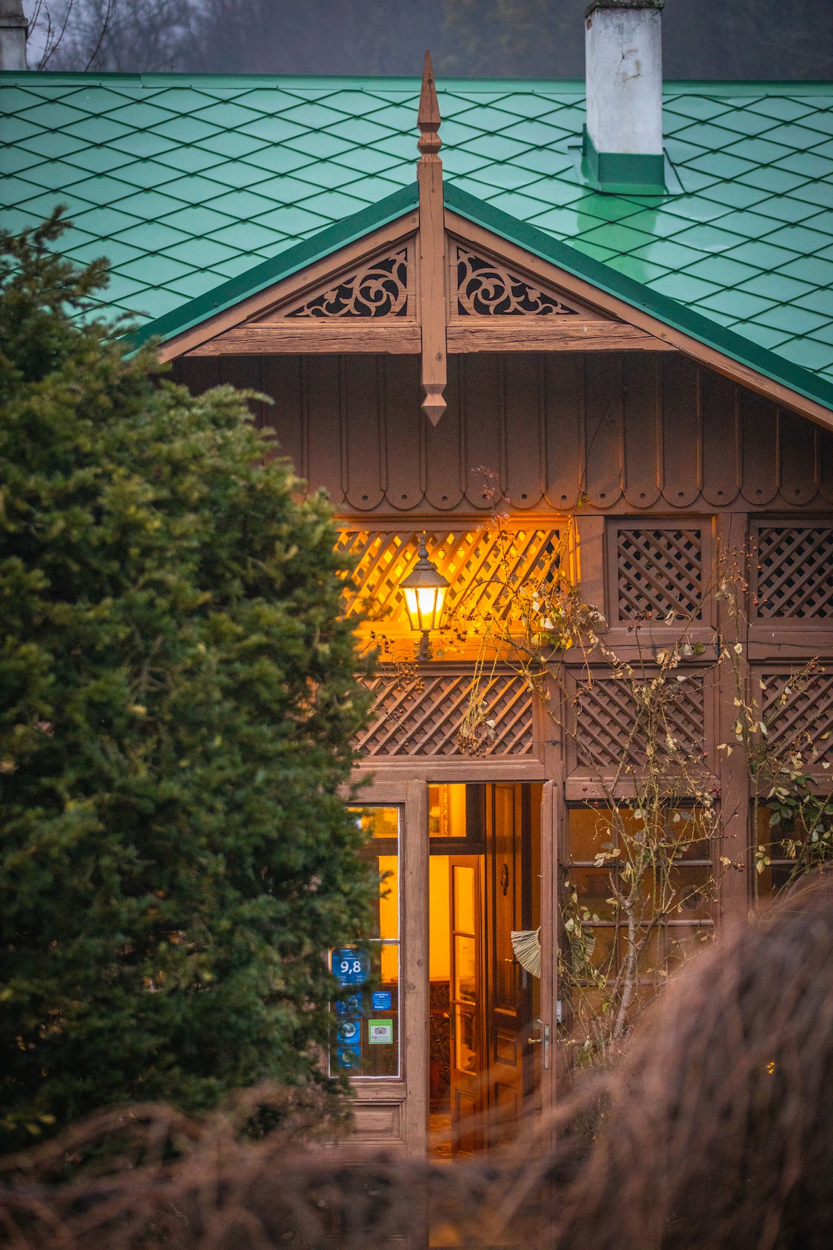 The cottage entrance is framed by green foliage and characterized by intricate wooden accents. A warm glow emanates from the lantern, illuminating the entrance, while a glimpse of the interior is visible through the doorway. The distinctive green roof adds to the cottage's charm.