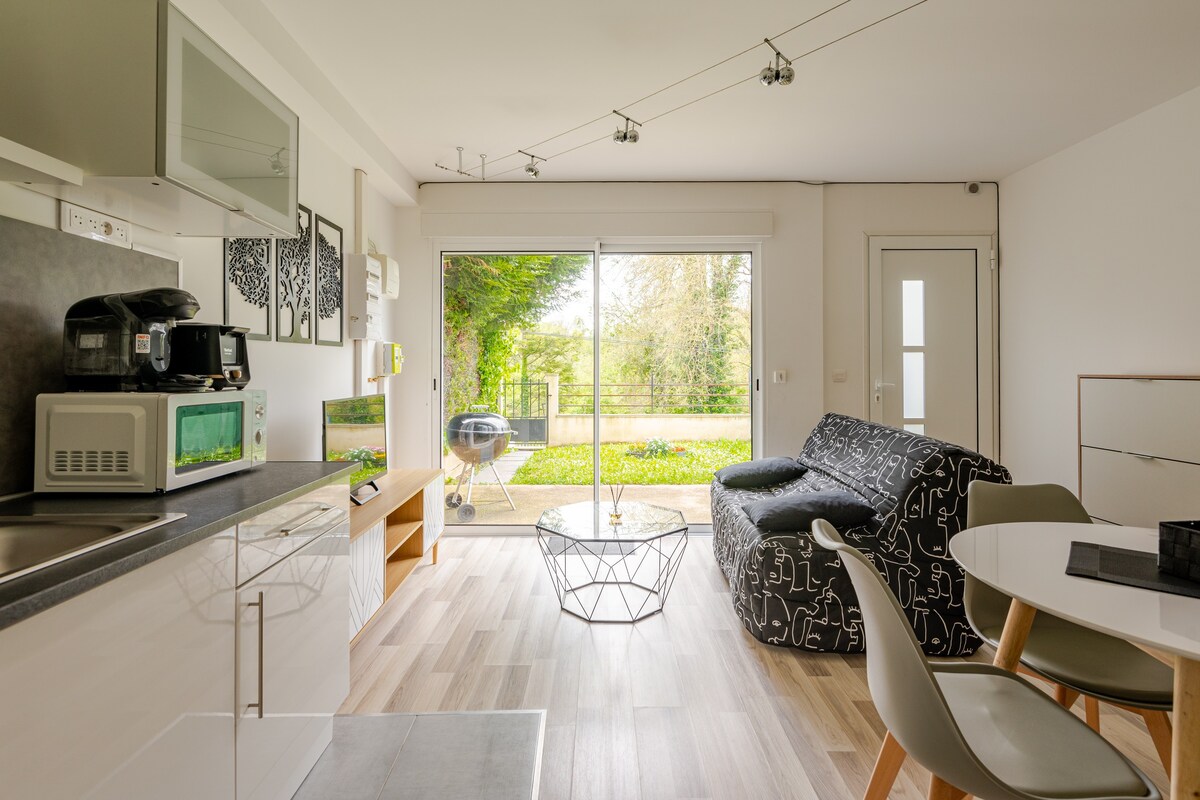 A modern living space includes a well-equipped kitchen area with a microwave and coffee machine. A dining table is set beside a cozy sofa, while large glass doors provide a view of the green outdoor space, enhancing the ambiance.
