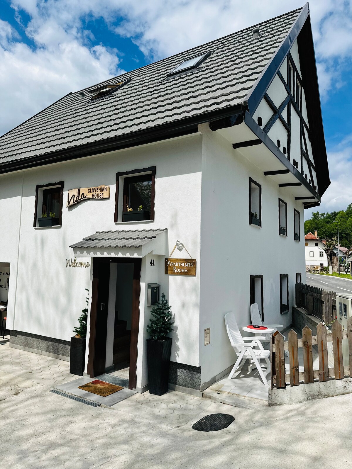 A traditional-style house features a white exterior with wooden accents and a sloped roof. The entrance is highlighted by a welcome sign and decorative potted plants, while a small white table and chair are positioned outside, inviting guests to sit and enjoy the serene surroundings.