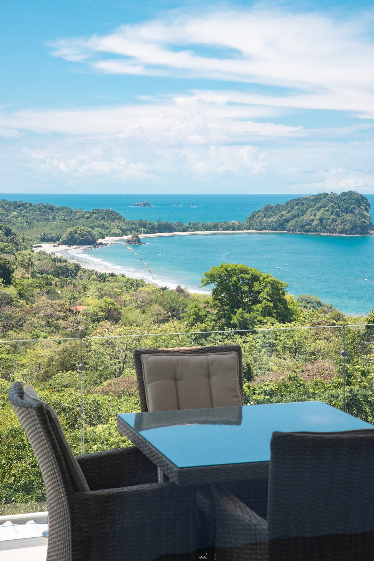 A dining table with four woven chairs is positioned on a private balcony, offering a clear view of the lush coastline and ocean. Vibrant greenery frames the scene, while the azure water reflects the bright sky dotted with fluffy clouds.