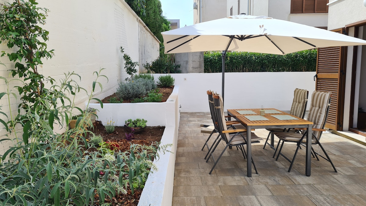 An outdoor dining area features a large umbrella above a wooden table surrounded by six chairs. The space is bordered by a well-maintained garden with various plants, offering a private setting for relaxation. The ground is paved with light-colored tiles.