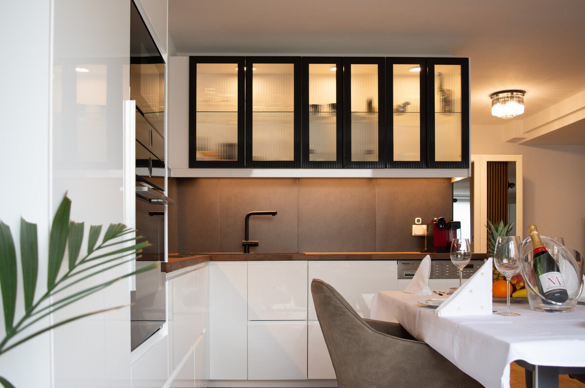 A modern and fully equipped kitchen is depicted, featuring sleek white cabinetry and contrasting black glass doors. A minimalist faucet and a stylish dark countertop are visible. A dining table is set nearby, complemented by elegant glassware, suggesting a space ideal for meal preparation and dining.