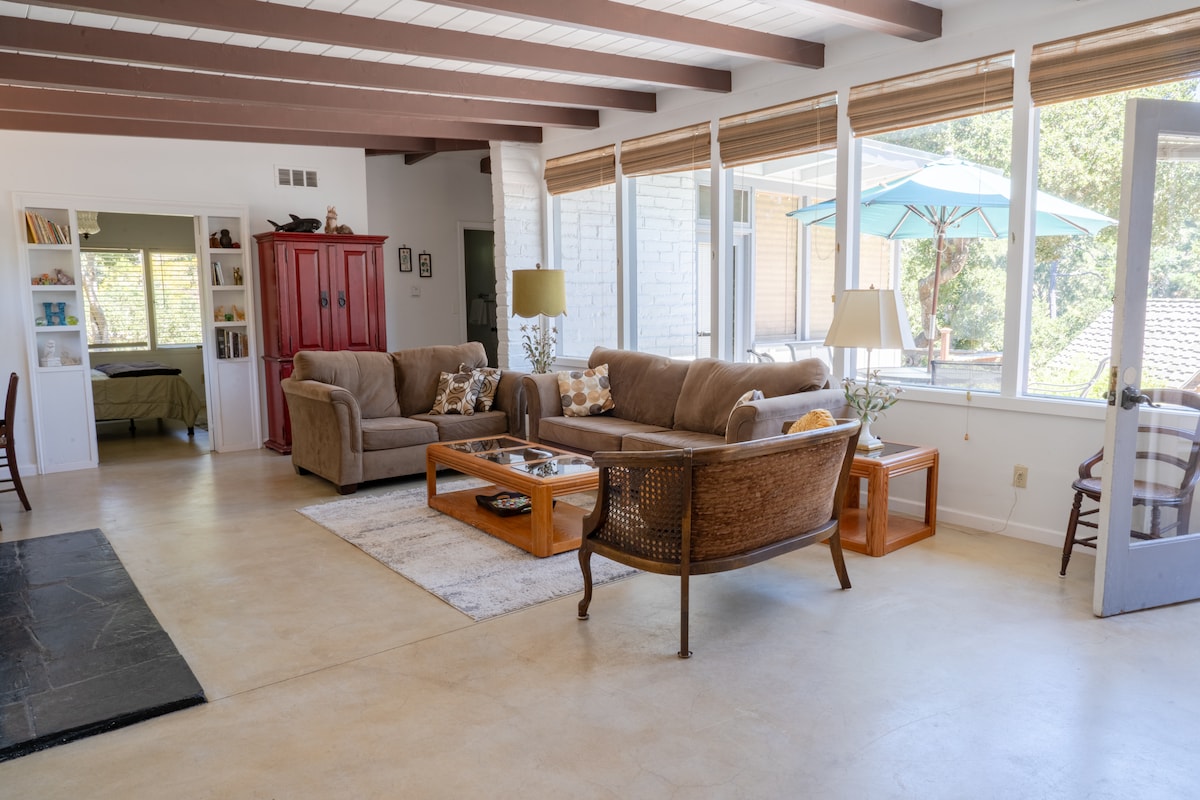 A spacious living area features large windows allowing natural light to illuminate the space. Comfortable seating includes a brown sofa, a woven lounge chair, and a wooden coffee table. A red cabinet adds a pop of color, while an outdoor patio is visible through the glass doors.