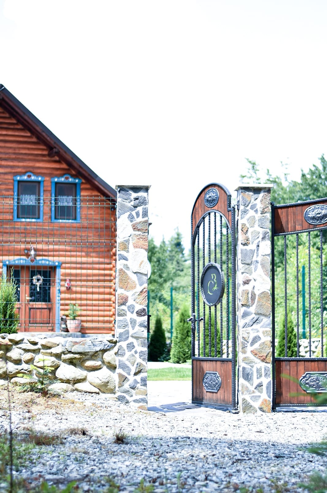 An inviting entrance gate framed by stone pillars opens to a charming wooden house. The house features a welcoming front door adorned with simple decorations. Surrounding greenery enhances the peaceful ambiance, offering a glimpse of the serene outdoor space.