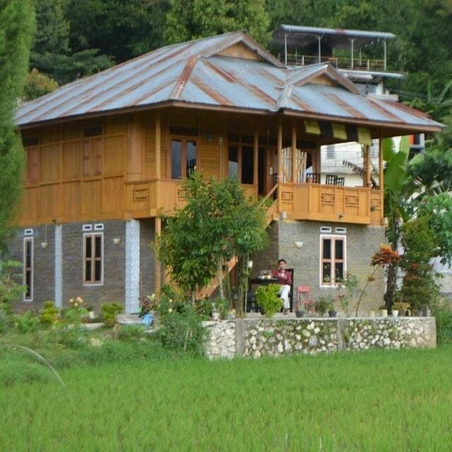 A two-story wooden house with a sloped roof is surrounded by lush greenery and rice fields. The exterior showcases a mix of wood and stone elements, with balconies enhancing the view of the landscape. A bicycle is nearby, indicating accessibility to the countryside.