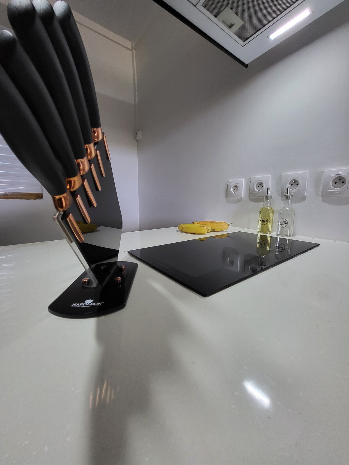 A modern kitchen counter is displayed, featuring a set of knives on a sleek stand. An elegant glass cooktop is visible alongside two decorative bottles. The countertop surface appears smooth and light in color, adding to the contemporary design.