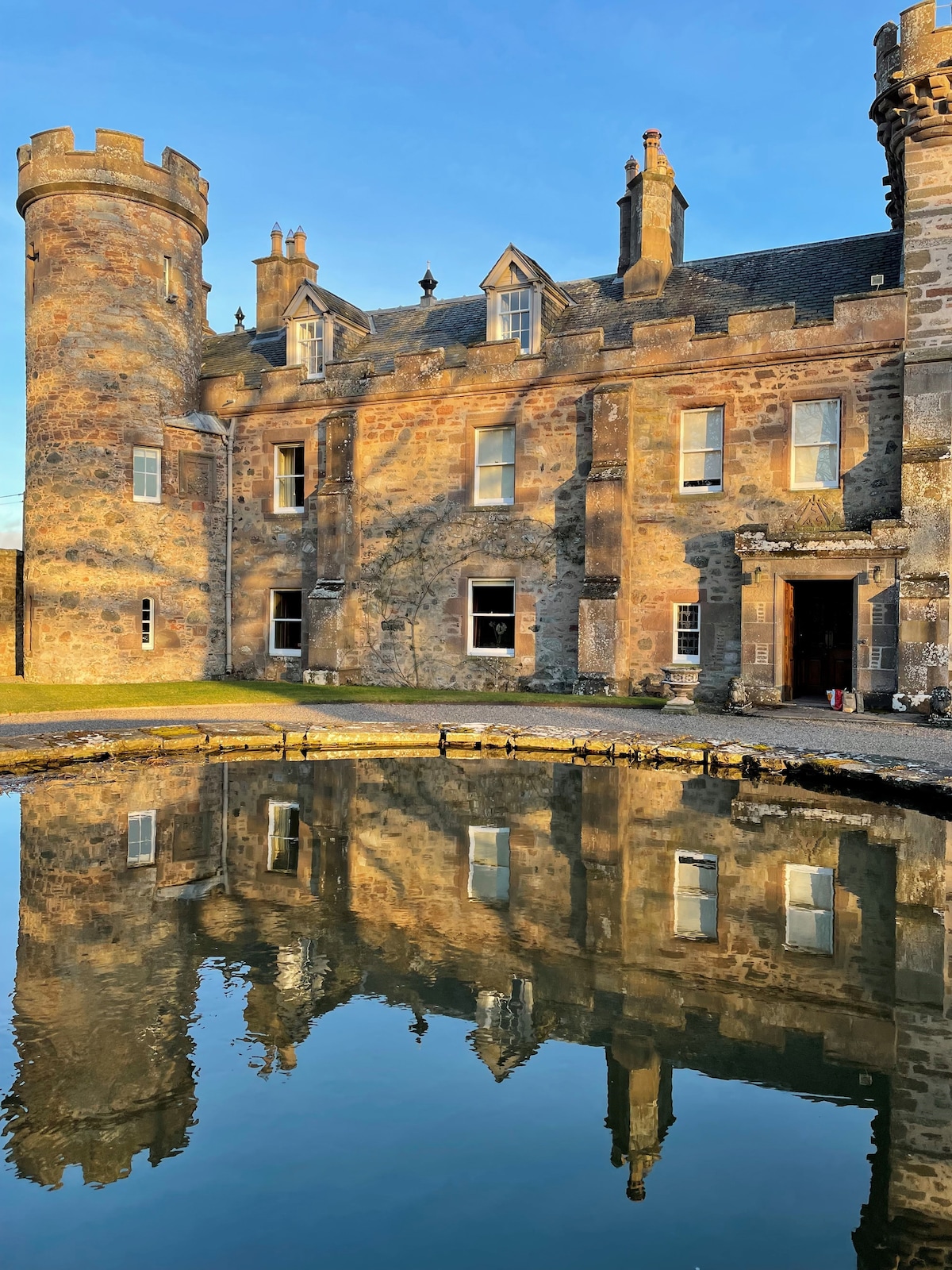 The exterior of a historic castle is captured at sunset, with stone walls displaying intricate architectural details. A tranquil pond reflects the structure, featuring a turret and a large entrance. The surrounding green space adds a touch of nature, enhancing the serene environment.
