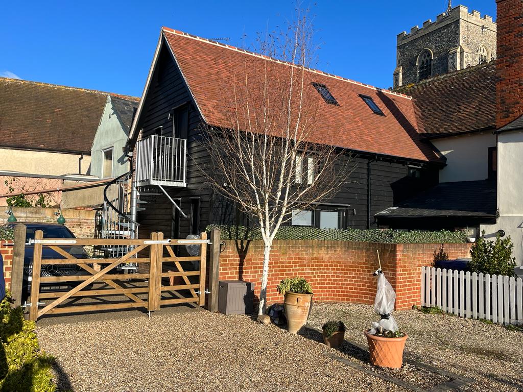 The exterior of The Hayloft is shown, featuring a black timber building with a red-tiled roof. A gravel driveway leads up to a wooden gate, and a small tree in a pot adds a touch of greenery. The church tower is visible in the background.