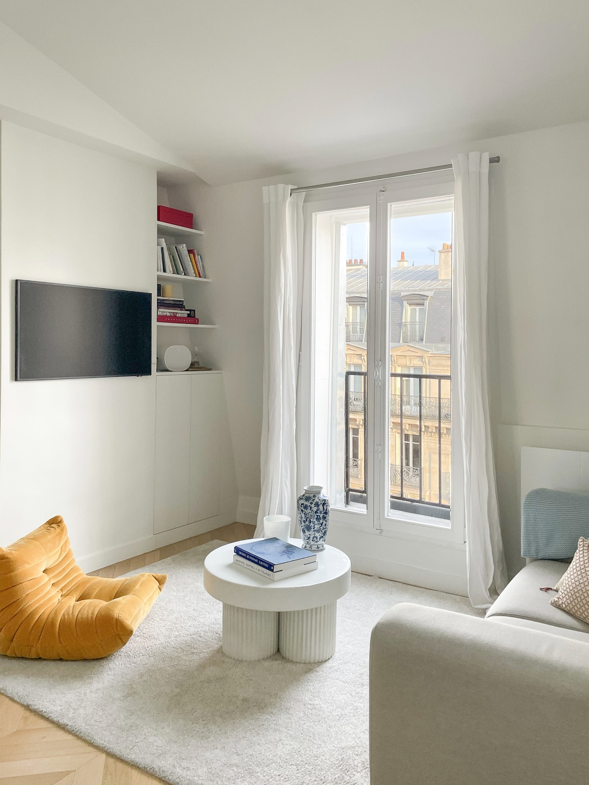 A living area is revealed, featuring a light color palette. A cozy yellow pouf and a minimalist white coffee table occupy the center. A wall-mounted television is visible, with built-in shelving containing books. Natural light streams through wide windows overlooking Parisian rooftops.