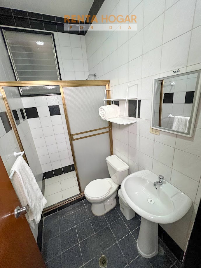 A bathroom is displayed, featuring a shower area with glass doors and tiled walls in black and white. A white toilet and sink are positioned adjacent to each other. A neatly folded towel rests on a shelf, while a mirror is mounted on the wall.