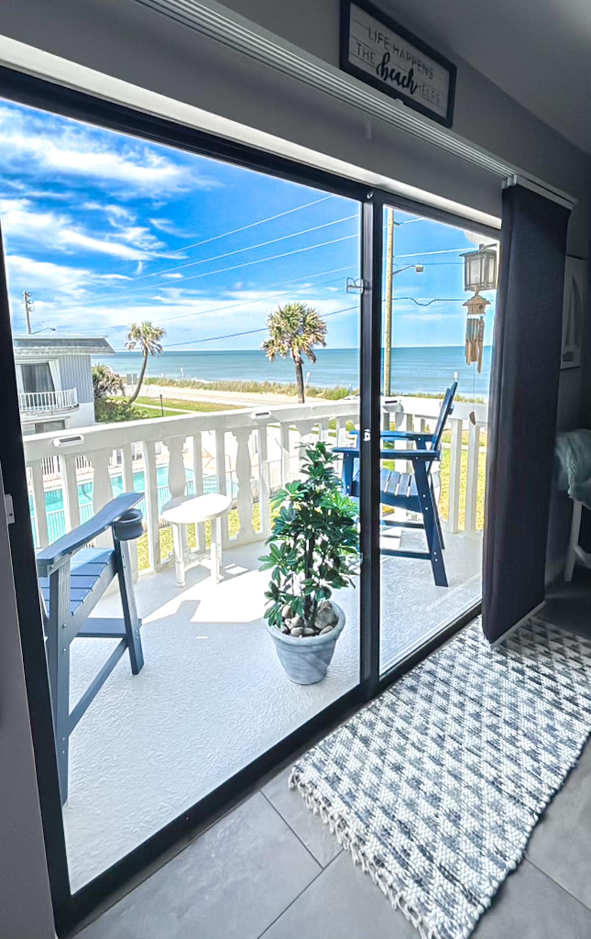 A sliding glass door opens to a private balcony with ocean views. The balcony is furnished with blue chairs and a small table, alongside a potted plant. A patterned rug is visible on the interior floor, providing a smooth transition to the outdoor space.