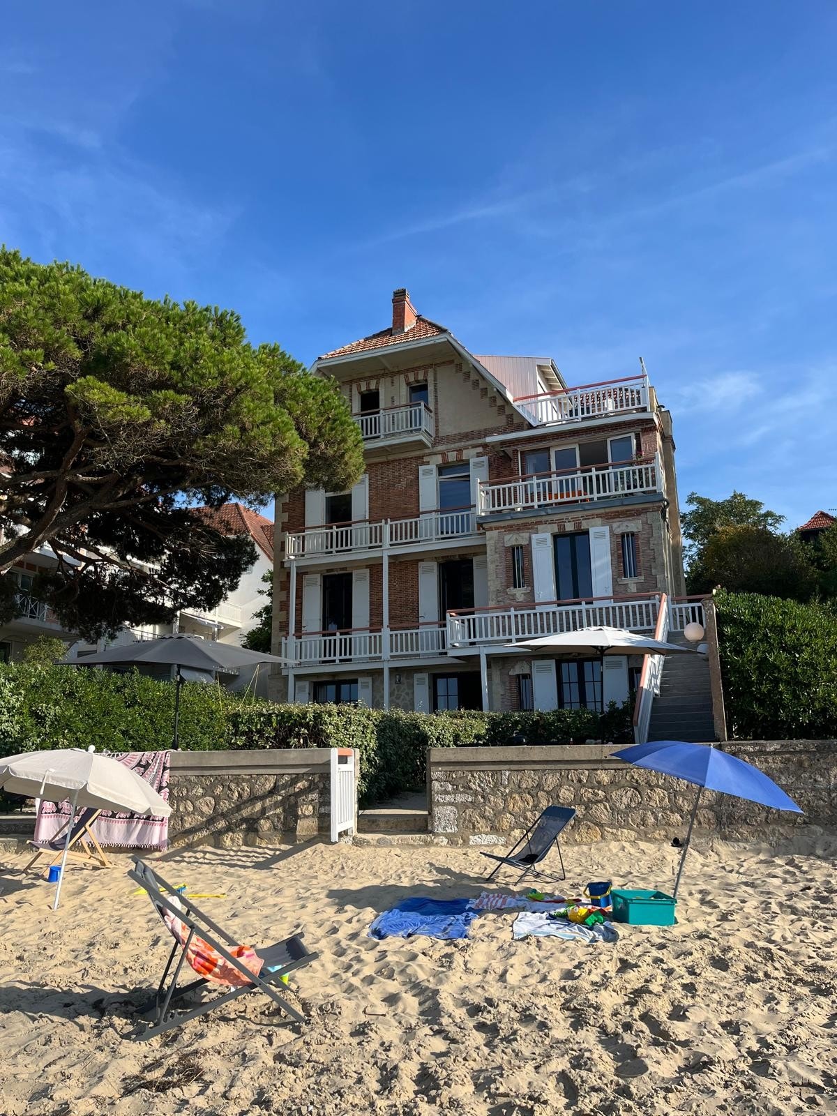 A charming three-story villa is situated near the beach, showcasing multiple balconies framed by lush greenery. Chairs and umbrellas are set up on the sandy beach in the foreground, while the building's architectural features highlight its traditional style.