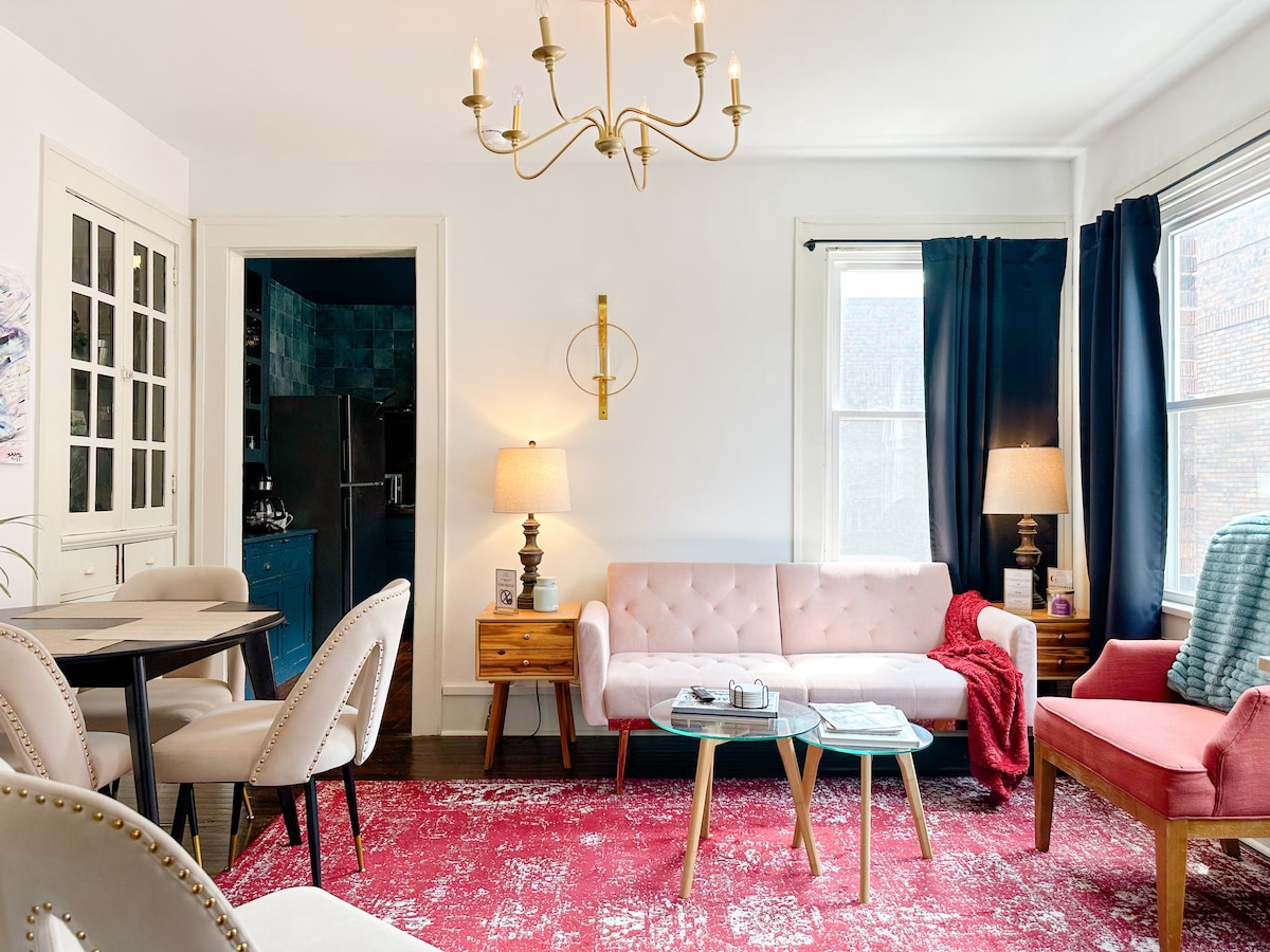 A bright living area features a pink couch paired with a glass coffee table, surrounded by a dining table and chairs. Natural light streams through large windows, highlighting a stylish chandelier. A textured red rug adds warmth, while neutral walls create an inviting backdrop.