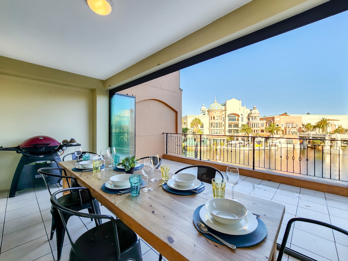 A balcony dining area is presented featuring a wooden table set for six, complete with dishes, glasses, and cutlery. A gas grill is positioned nearby, and large sliding doors open to stunning canal views, enhancing the atmosphere for outdoor dining.