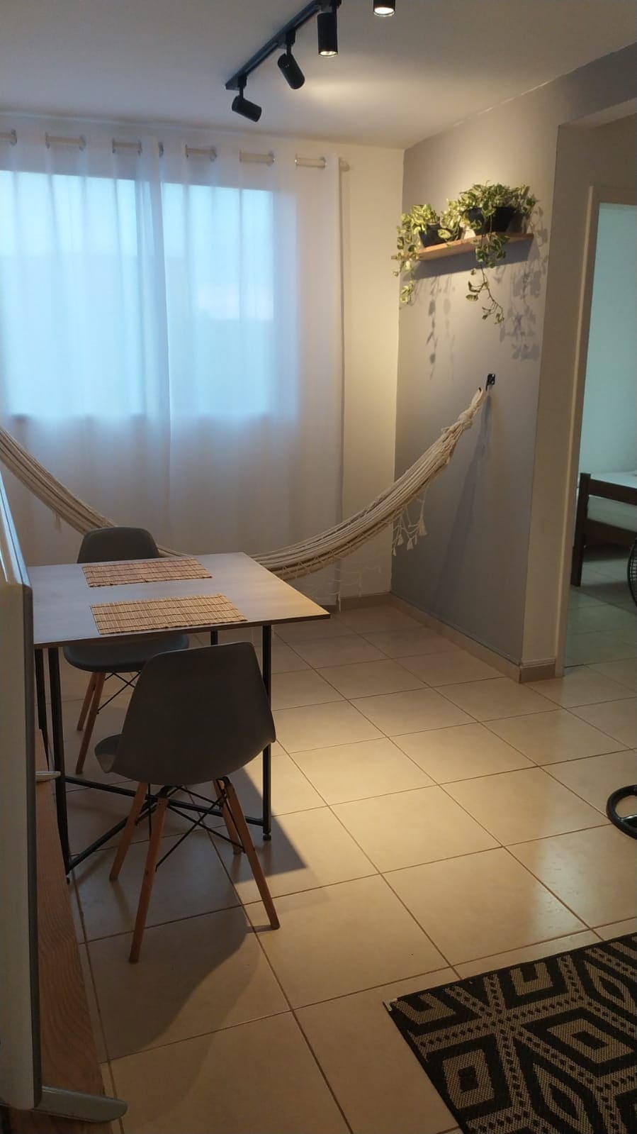 A dining area is furnished with a simple table and four chairs, accompanied by a hammock gracefully suspended between the walls. Natural light filters through sheer white curtains, illuminating the light-colored tile floor and enhancing the minimalist décor.